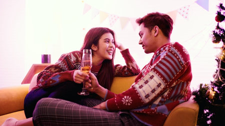 Candid couple in cozy sweaters toast champagne on a sofa by a decorated Christmas tree, sharing...
