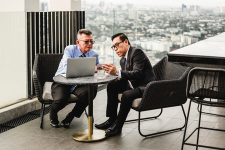 Two professionals in suits collaborate over laptop on rooftop terrace with city view. Outdoor business meeting blends planning, leadership, sales, deal, conference, networking, and partnership.の写真素材