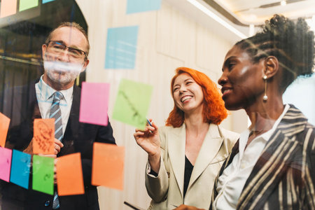 Diverse coworkers brainstorming with colorful sticky notes on a glass wall in a modern office, smiling and collaborating during a creative planning meeting.の写真素材