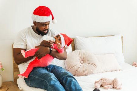 Dad in a Santa hat feeds a baby with a bottle on a cozy bed at home, both in festive outfits. Warm, intimate family moment celebrating the holidays.の写真素材