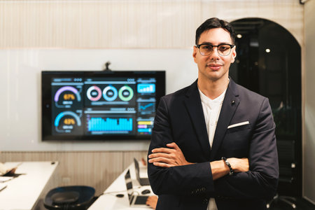 Confident businessman in a modern office stands with arms crossed beside a data dashboard display, conveying leadership, strategy, and digital transformation.の写真素材