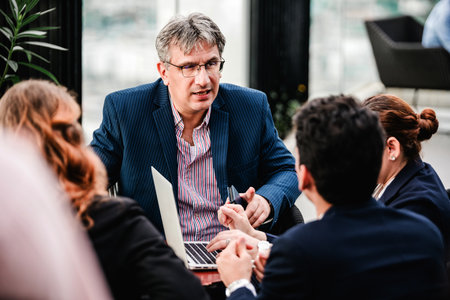 Senior executive leads a business meeting with colleagues, discussing strategy around a laptop in a...