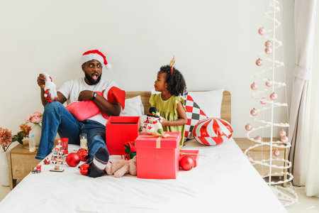 Father and daughters in festive hats exchange Christmas gifts on a cozy bed. Surrounded by ornaments and presents, the joyful holiday scene captures family celebration, happiness, and togetherness.の写真素材