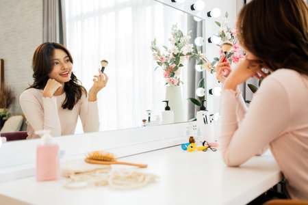 Smiling woman applying makeup at a bright vanity, looking into a large mirror with cosmetics, flowers, and accessories on the table in a sunlit modern bedroom.の写真素材