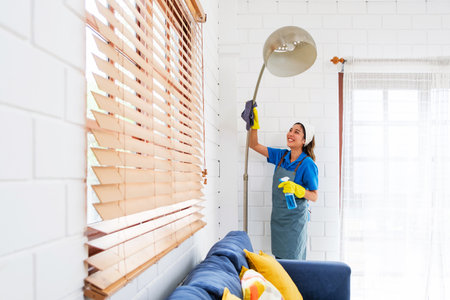 Smiling house cleaner dusts a tall floor lamp in a bright living room, wearing gloves and apron...