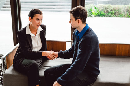 Two professionals sit in a modern lobby, shaking hands after a meeting, symbolizing partnership, agreement, and trust in a corporate setting.の写真素材