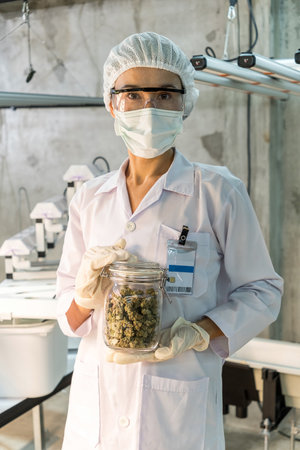 Caucasian lab technician in protective gear holding a jarred sample in a sterile facility, demonstrating quality control and safety procedures with mask, gloves, goggles, and hairnet.の写真素材