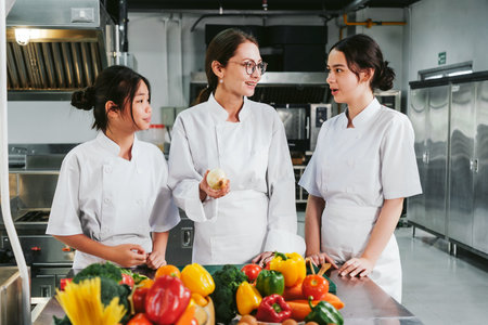 Three chefs in white uniforms discuss prep in a professional commercial kitchen, surrounded by colorful fresh vegetables on a stainless counter, conveying teamwork, training, and culinary planning.の写真素材