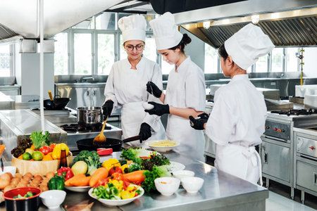 Three chefs in white uniforms and hats cook in a professional stainless steel commercial kitchen, preparing dishes at the stove with fresh vegetables and ingredients arranged on the counter.の写真素材