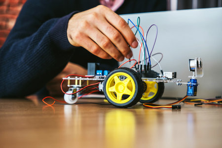 Close-up of a person assembling a small wheeled robot beside a laptop, connecting wires for a robotics project, illustrating STEM education, engineering, and prototyping.の写真素材