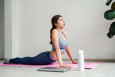 Asian young woman practicing cobra pose on a pink yoga mat at home, breathing deeply beside a laptop and water bottle in a bright, minimalist room with a plant. New year resolution, treat yourself.の写真素材