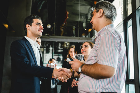 Professionals at a networking event shake hands while others chat in a modern indoor venue.の写真素材