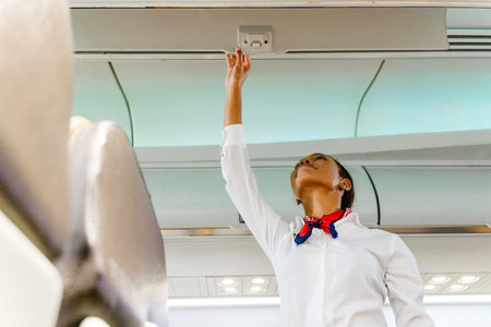 Flight attendant in uniform reaches to adjust an overhead panel inside a modern airplane cabin during preflight checks. Inflight cabin airline service business conceptの写真素材