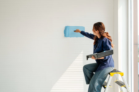 Person on a step ladder painting a white interior wall with a blue roller, starting a home makeover in natural daylight.の写真素材
