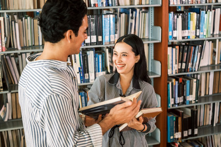 Two students laugh and chat with books in hand in a library aisle, surrounded by shelvesâcapturing campus study life, learning, literature exploration, seminar prep, and academic resources.の写真素材