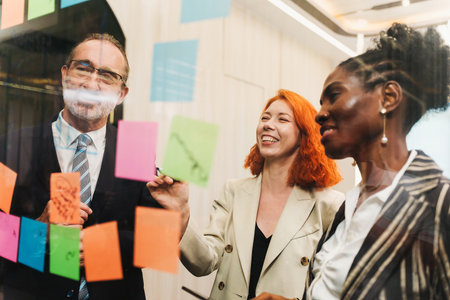 Diverse professionals smiling and collaborating during a brainstorming session, using colorful sticky notes on a glass wall in a bright modern office, showing teamwork and creative planning.の写真素材