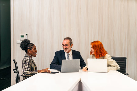 Three business professionals meet around a modern office table, each working on laptops and actively engaging in discussion, reflecting teamwork, collaboration, and corporate strategy.の写真素材