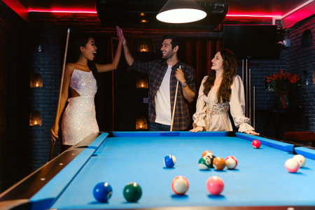 Three friends cheer around a pool table in a stylish, dimly lit billiards lounge, enjoying a night of laughter, competition, and connection in a vibrant nightlife setting.の写真素材