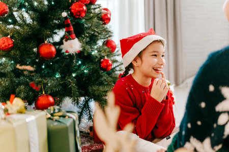 Cheerful girl daughter in a Santa hat celebrates beside a decorated Christmas tree, surrounded by wrapped gifts in a cozy living room. Indoors Christmas family conceptの写真素材