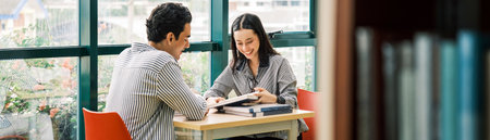 An Asian woman and Caucasian man sit by a large window in a bright library or office, engaged in...