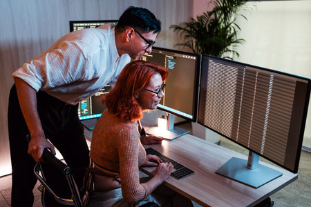 Two people focus on coding and analyzing data across multiple computer monitors in a bright office,...