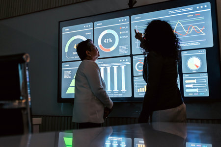 Two professionals analyze data on a large touchscreen, pointing at colorful dashboards and graphs in...