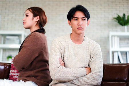 A young couple sits back-to-back on a couch with arms crossed, conveying relationship tension and emotional disconnect in a home setting â a relatable scene of disagreement in modern life.の写真素材