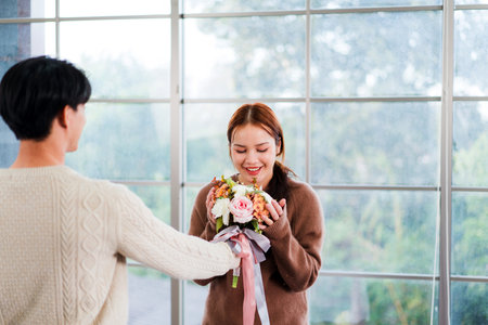 A man surprises a smiling woman with a bouquet of flowers in a bright indoor space by a large window, capturing a tender moment of romance, joy, and affection in a heartfelt relationship.の写真素材