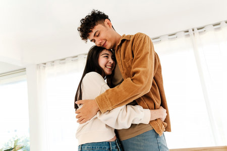 A young couple shares a joyful embrace in a bright room, smiling warmly and enjoying a happy moment together near large windows with natural light. Valentines Day conceptの写真素材