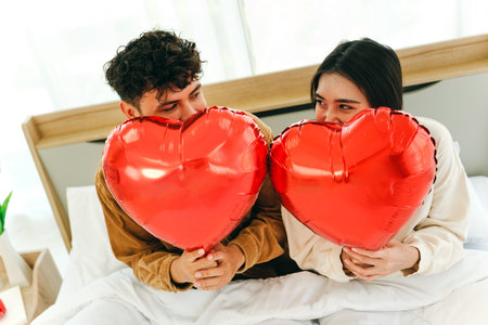 A loving couple holds heart-shaped balloons and playfully gazes at each other in a cozy setting,...
