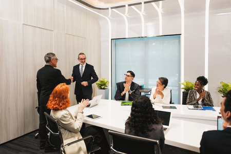 Diverse team in a modern conference room applauds as two colleagues shake hands after a successful...
