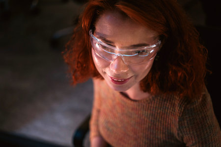 Close-up of a person with red hair wearing clear safety glasses, lit indoors, suggesting a lab or...