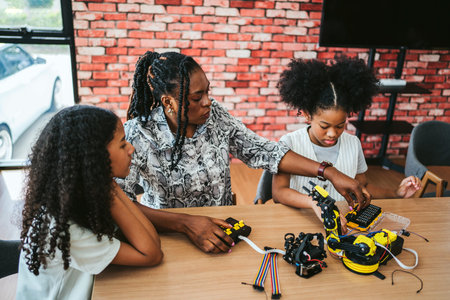 Teacher guides two girls building a robotic arm kit in a classroom makerspace, using electronics and...