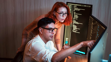 Two software developers collaborate at dual monitors, reviewing code in a dim office; one points at...