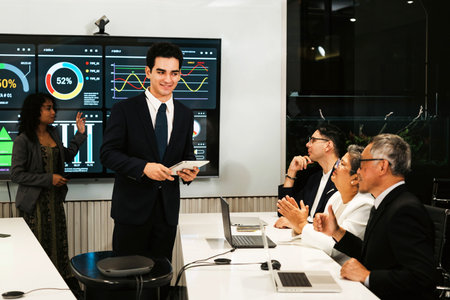 Professionals in a modern conference room review analytics dashboards during a presentation, as the presenter stands with a tablet and colleagues discuss and applaud.