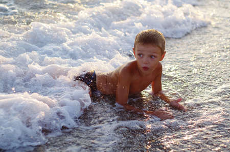 A happy boy is playing in the waves on the beach. Cheerful boy bathes in the sea waves at sunsetの写真素材