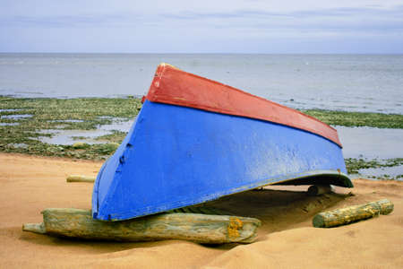 A blue-red boat lies upside down on logs on the sandy seashore.の写真素材