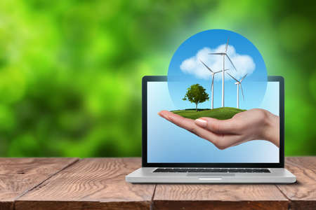 Wind turbines on meadow with tree holds in woman hand against blue sky and clouds in open laptop on wooden table. Worldwide Green energy conceptの写真素材