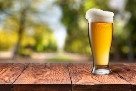 Beer in glass on wooden table with blurred green summer leaves on natural background with bokehの写真素材