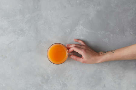 Female hand holding the glass of orange juice on gray stone background, flat layの写真素材