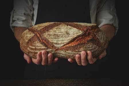 baker's hands hold a fresh oval bread on the background of a black apronの写真素材