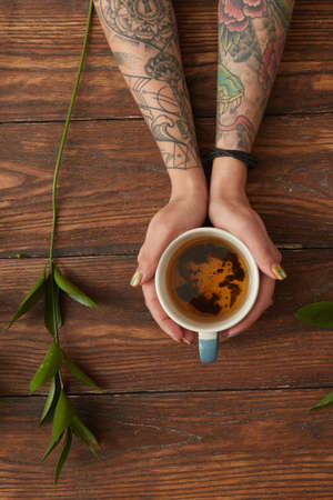 Female hands with tattoos holding a cup of fragrant tea on a wooden table near a green branch flat...