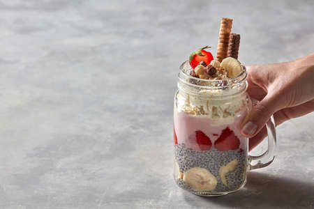 Glass with a layered breakfast from yogurt, strawberry, chia pudding with a banana in the female...