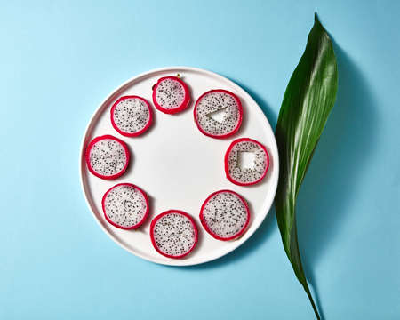 Round pieces of Pitahaya in a white plate and green leaf on a blue paper background. Top viewの写真素材