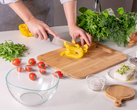 A womans hand slices a yellow pepper on a wooden board on a white kitchen table. Preparation of...