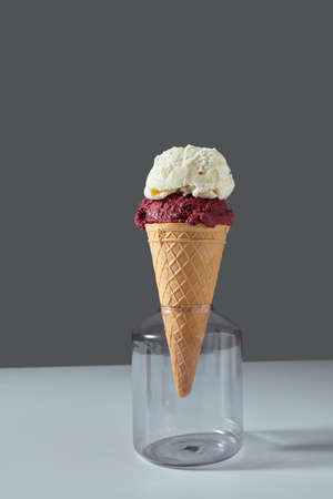 A balls of berry and vanilla ice cream in a waffle cone stands in a glass jar on a gray background...