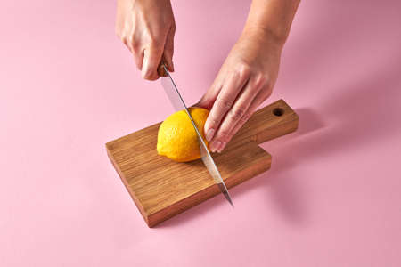 Citrus fruits on cutting board. Female hands cutting a yellow ripe lemon on half on a wooden board on a pink background. Concept vegetarian food.の写真素材