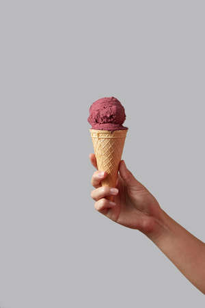 A female hand holds red berry ice cream in a waffle cone on a light gray background.
