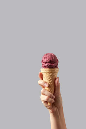 A womans hand holds a waffle cone with red cherry sorbet on a light gray background.の写真素材
