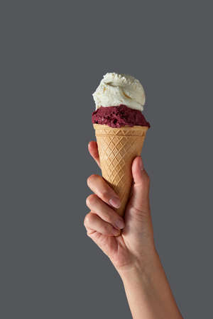 A female hand holds red berry gelato and white milk ice cream in a waffle cone on a gray background....